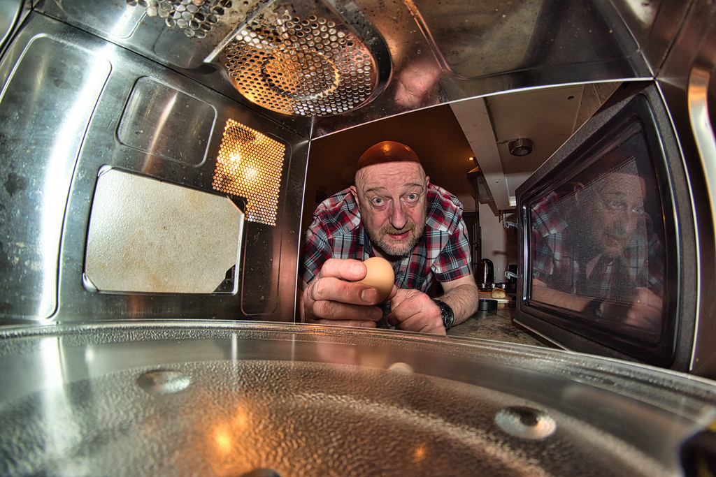 A man holding an egg is leaning into a microwave, towards the camera which is positioned inside